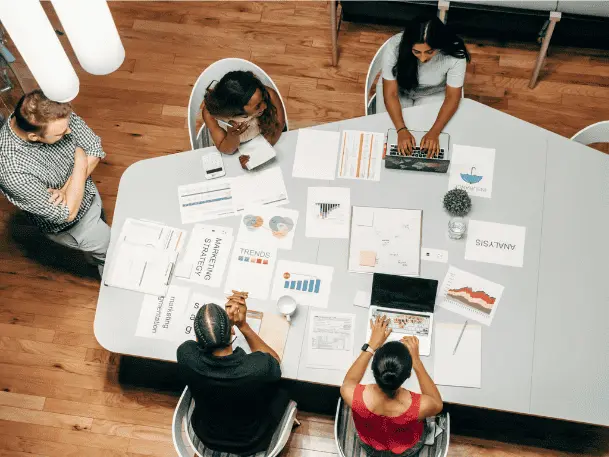 Overhead view of a team working around a table covered in printed charts and laptops
