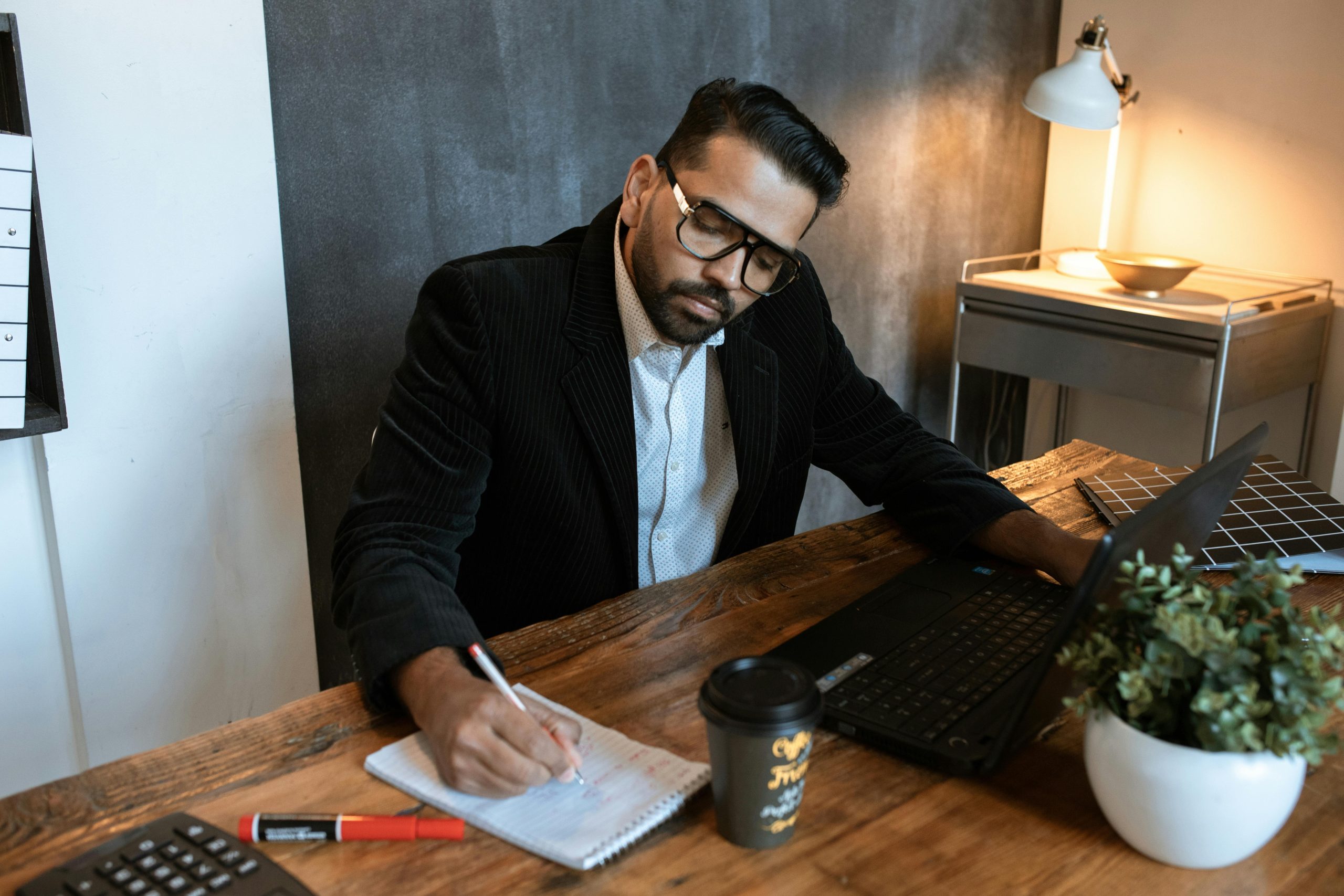 Man with glasses and a suit sitting at his table writing notes while working on a computer